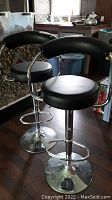 Two black high bar stools with padded seats and backs, chrome metal frames with footrests and rotating bases seen from front angle.