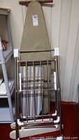 Large beige ironing board with cover and frame with smaller blue and white striped ironing board leaning against the wall behind a folded metal drying rack with white plastic joints.