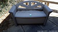 Full front view of the gray Rubbermaid patio bench storage unit on an outdoor patio surface, showing backrest, armrests, and seat surface with some debris.