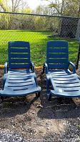 Pair of blue plastic patio lounge chairs side by side outdoors, showing slatted wide seats and adjustable backrests with armrests.