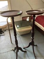Wide shot showing both wooden plant stands with round tops, spindle center columns, and three legs each in a room corner.