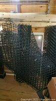 Photo showing three rolls of black plastic fencing with diamond mesh pattern, indoor storage on wooden floor near wooden wall.