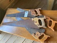 Four rusty finishing hand saws with wooden handles placed on a wooden surface, all blades facing the same direction, showing handles and blade conditions.