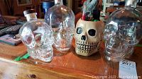 Photo of three clear glass skull-shaped liquor bottles and one beige ceramic skull pen holder with pens and pins on a wooden surface.