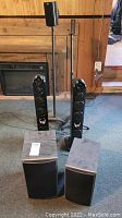 Two tall Athena Audition Series tower speakers on the floor with two separate tall black speaker stands behind them, all placed in front of a fireplace in a carpeted room.