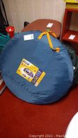 Blue fabric carrying case for Montague pop-up tent with orange straps and attached product label.