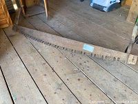Full view of the vintage two-person cross-cut saw lying on wooden floorboards, showing length and condition.