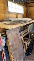Wide view showing stacked wood planks and plywood leaning and stored inside workshop near window.