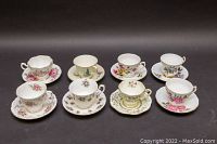 Photo of eight bone china teacup and saucer sets arranged on a black backdrop, showing floral patterns and gold trim on each set.