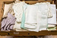 Box of assorted linen tablecloths and napkins showing various folded items in white, cream, lavender and mint green, some with lace or embroidery.