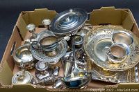 Top-down view of all items in cardboard box displaying shuffled arrangement including trays, candleholders, creamer, salt & pepper shakers, ladle and other silver plated serving pieces.