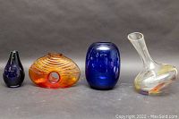 Four mid-century modern blown glass items on neutral background; two cobalt blue vases, an orange vase with hole and textured design, and a clear decanter with orange and purple swirls.