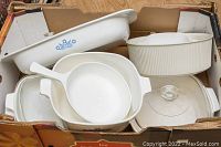 Box containing assorted white casserole dishes including several with blue floral CorningWare Cornflower pattern and ribbed oval dish, some glass lids