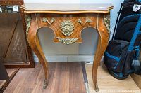 Front view of demi-lune table showing curved legs, brass accents shaped like a face and floral motifs, marquetry top, and aged wood surface with water damage.