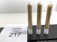 Front view of three small transparent tubes each filled with 24k gold flakes approximately three quarters full, placed upright on a black surface alongside a maxsold number card.