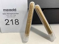 Two clear plastic test tubes filled with gold flakes shown upright on white surface with auction lot sign in background.