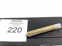 Clear plastic test tube containing fine gold flakes seen on black surface with lot number tag.