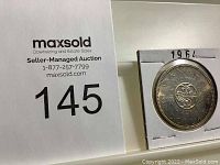 Coin in holder alongside Maxsold lot number 145 card showing the coin for auction listing.