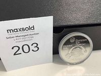 Photo shows silver coin in clear protective capsule with MaxSold auction card.