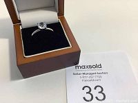 Silver solitaire ring set in wooden box with black insert and MaxSold auction card in background.