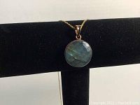 Close-up photo showing pendant with blue-green faceted stone on gold-colored chain on black display stand