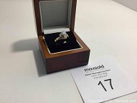 Ring inside a wooden jewelry box with black velvet interior, showing the large clear center stone and smaller side stones.