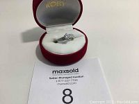 Silver ring with central round clear stone and smaller stones on the band placed inside a red KOBY jewelry box on white cushioning.