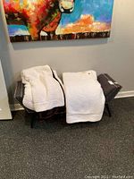 Photo of gray tufted fabric bench with studded trim and two white plush blankets over it under a colorful painting.