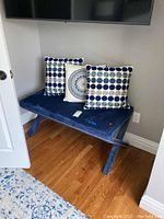 Blue velvet bedside bench with three blue and white patterned throw pillows placed on it, shown angled in a corner of the room on hardwood floor with blue rug nearby.