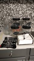 Four glass canisters with black lids and chalkboard labels with contents inside, five plastic scoops, white napkins in metal holder, black metal sign on grey surface.