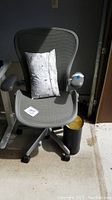Front view of gray mesh office chair with lumbar pillow on seat and black metal trash bin at base.