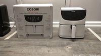 Boxed and unboxed Cosori 5.8 quart air fryers of model CP158-AF shown side-by-side on a floor.