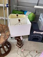Two modern style table lamps with rectangular fabric shades and wooden bases placed on a concrete floor surrounded by various items including a dog house and shelving units.