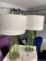 Pair of tall green glass lamps with white drum shades, showing the overall style and design of the lamps in a room setting with background elements like chairs and a bicycle.