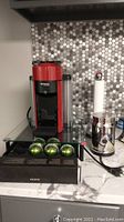 Red and black Nespresso VertuoLine coffee machine on countertop with Melozio coffee pods in opened box and 5 Alto Intenso pods arranged in front, a stainless steel coffee grinder, and white paper towel holder visible.
