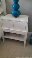 Photo of one white wooden nightstand with a blue lamp on top showing drawer and open lower shelf.