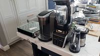 Wide shot showing Ninja blender and Keurig coffee maker side by side on kitchen counter with assorted coffee pods tray.