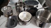 Overview of assorted pots and pans including large stock pot, stackable steamers, cast iron skillet, and decorative trays on kitchen counter.