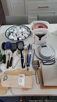 Kitchen utensils including spatulas, tongs, slotted spoon, peeler, whisk, white ceramic egg cups on floral tray; digital and analog scales; metal trivets; wooden cutting boards with blue loops
