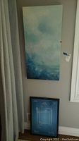 Blue and white abstract painting hanging on wall above framed blueprint print leaning against wall