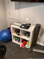 Fitness equipment including Fitbit smart scale, AKG headphones, dumbbells, exercise ball, yoga mats, resistance bands, and weights placed around a white cube storage unit.