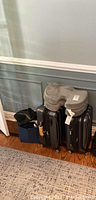 Two black Delsey hard-case roller suitcases side by side with one ComfiLife gray travel pillow on top. A blue container on left side holds gloves and scarves.