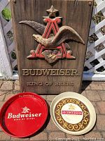 Full view of plastic Budweiser wall hanging and two Budweiser trays (red metal and white plastic) arranged outdoors on paved ground.