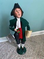 Front view of Byers Choice Williamsburg caroler figurine, holding lantern and music sheet, standing on green base.