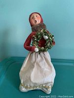 Full view of Byers Choice Victorian adult female caroler figurine holding wreath, wearing white skirt with gold trim, red top, fur scarf, and bonnet.