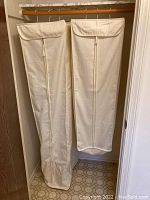 Two off-white cotton hanging clothes bags hanging on wooden rod inside closet, zippered front, flap closure at top.