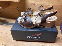 Side view of Dansko brown leather wedge sandals on top of a Dansko branded black box, highlighting wooden wedge heel and brown straps.