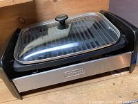 Front angled view of the DeLonghi indoor grill showing the glass lid with black knob and part of the grill body.