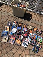 Wide shot of large plastic bin filled with DVDs and 31 DVDs laid out on ground showing variety of movie titles and cases.