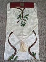Full view of Lenox Christmas table runner folded, showing holly and gold ribbon designs with tassel ends on white textured fabric.